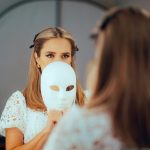 A woman stands in front of a mirror looking at herself. The lower part of her face is obscured by a plain white mask.